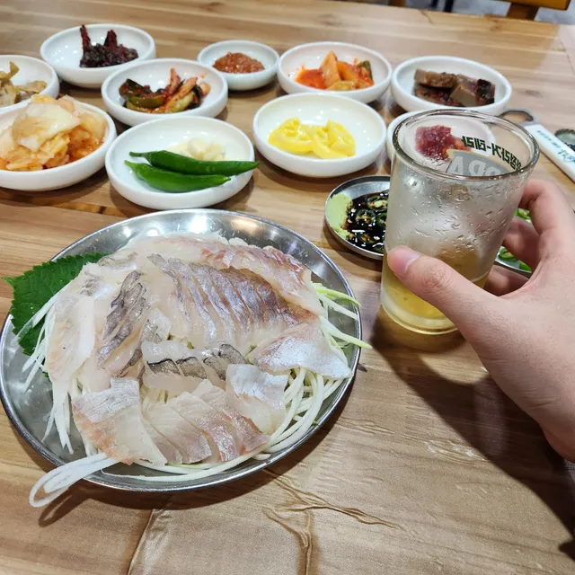 은쟁반에 담긴 생선회와 다양한 밑반찬, 맥주잔을 든 손이 있는 상차림
